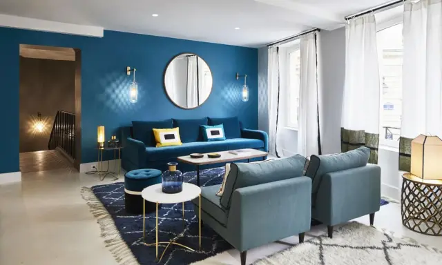 Salon moderne aux murs bleu canard, avec canapé velours, fauteuils assortis, tapis graphique et éclairage d'ambiance.