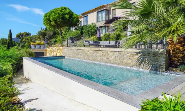 Belle piscine semi enterrée avec un habillage en pierre naturelle, entourée de verdure luxuriante et d'une villa moderne.