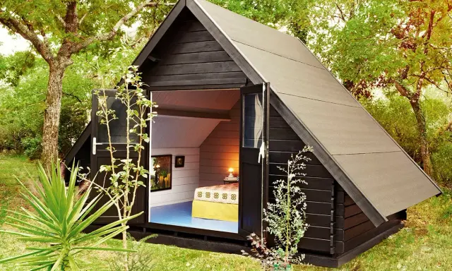 Aménagement abri de jardin : une cabane moderne en bois noir avec une porte ouverte révélant un lit douillet et des tableaux.