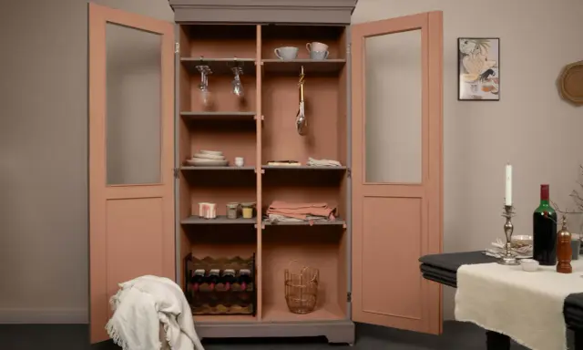 Armoire vitrée avec un relooking meuble 2 couleurs : gris anthracite et rose poudré. Elle contient vaisselle, verres, bouteilles de vin et paniers.