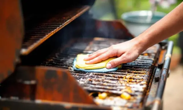 Main d'une personne nettoyant un barbecue très encrassé avec une éponge jaune et bleue.