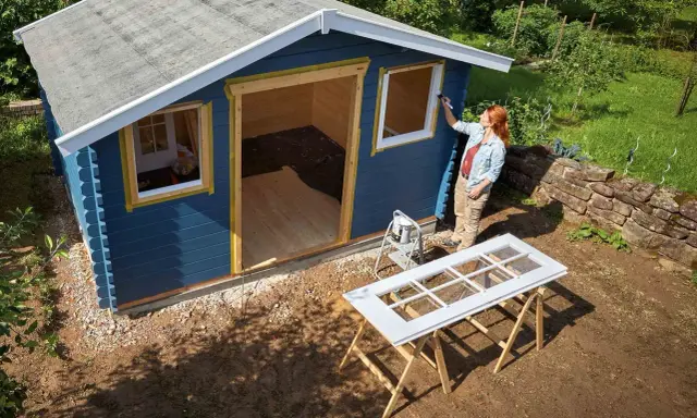 Une femme peint les contours des fenêtres d'une cabane de jardin bleue. Le toit de la cabane est en cours de finition.