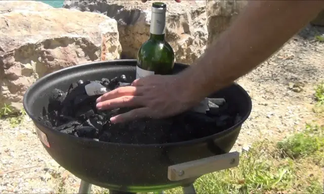 Main d'homme plaçant une bouteille de vin verte sur du charbon de bois dans un barbecue, étape pour comment allumer un barbecue.