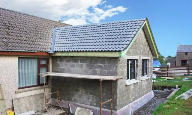 Agrandissement maison sur le côté en cours de construction, murs en parpaings, nouveau toit gris, échafaudage.