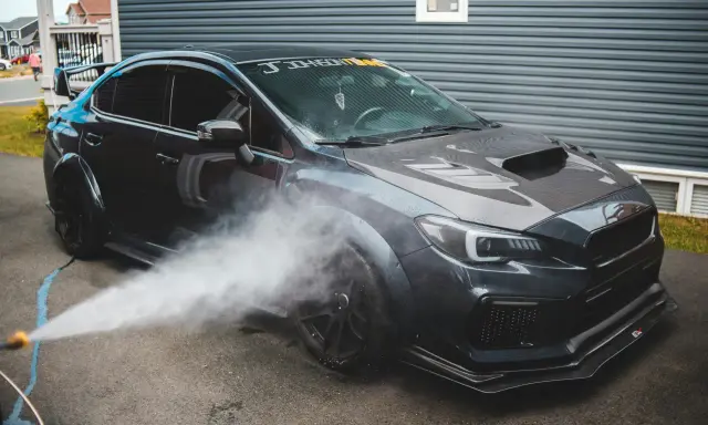 Nettoyage d'une voiture de sport sombre. Un jet d'eau est dirigé vers la roue avant, peut-être pour enlever mousse sur goudron vinaigre blanc.