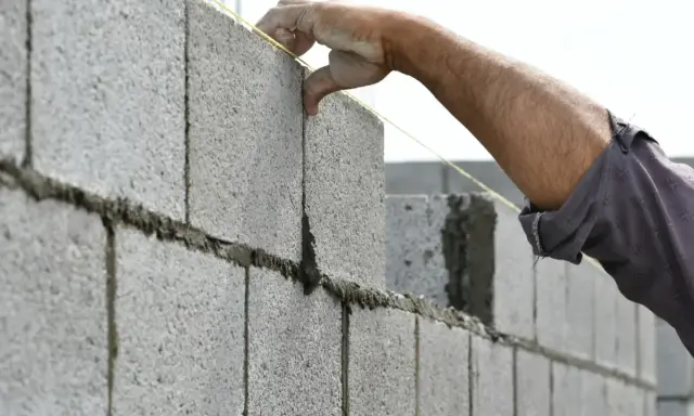 Main d'un maçon plaçant une brique sur un mur en parpaing, guidé par un fil à plomb.