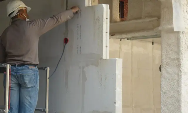 Un ouvrier en casque blanc travaille sur un mur en béton cellulaire, posant des blocs légers.