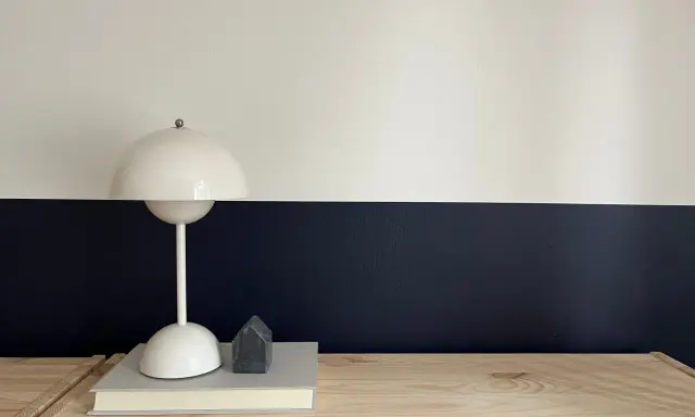 Lampe blanche sur un livre, devant un mur deux couleurs différentes, blanc et bleu nuit. Un petit objet gris en forme de maison est posé sur le livre.
