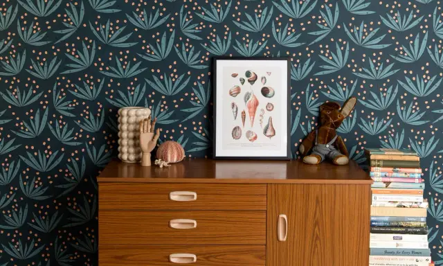 Idée motif peinture mur : papier peint botanique bleu nuit avec des feuilles stylisées et des baies roses. Un cadre de coquillages et une commode vintage complètent la scène.