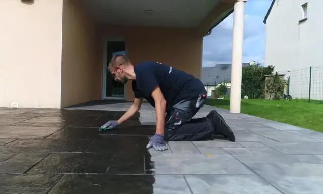 Un homme agenouillé effectue le nettoyage de dalles de terrasse avec une éponge verte, redonnant leur éclat à la pierre.