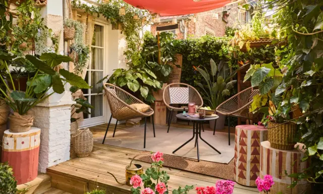 Aménager une petite terrasse sans se ruiner : trois fauteuils en rotin, une table basse, des plantes luxuriantes et une toile rouge créent un coin cosy et abordable.