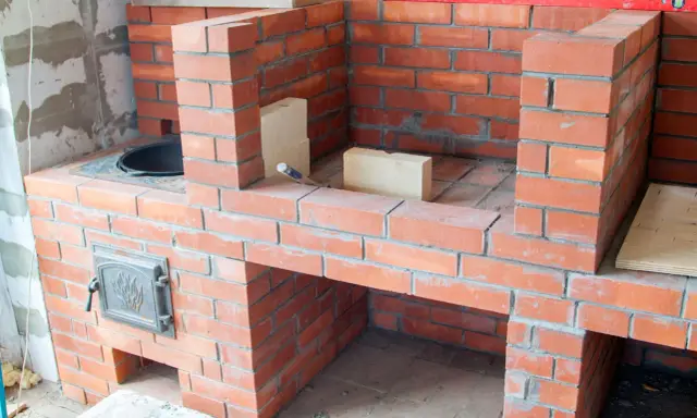 Construction d'un barbecue en brique, avec une porte en métal ornée et des briques claires pour la finition.