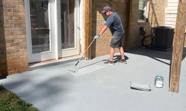 Homme appliquant une peinture sur mur béton extérieur avec un rouleau. Le sol de la terrasse est en cours de rénovation.