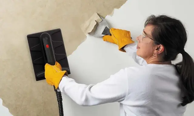 Une femme, portant des gants jaunes, utilise un appareil pour décoller du papier peint ancien d'un mur.