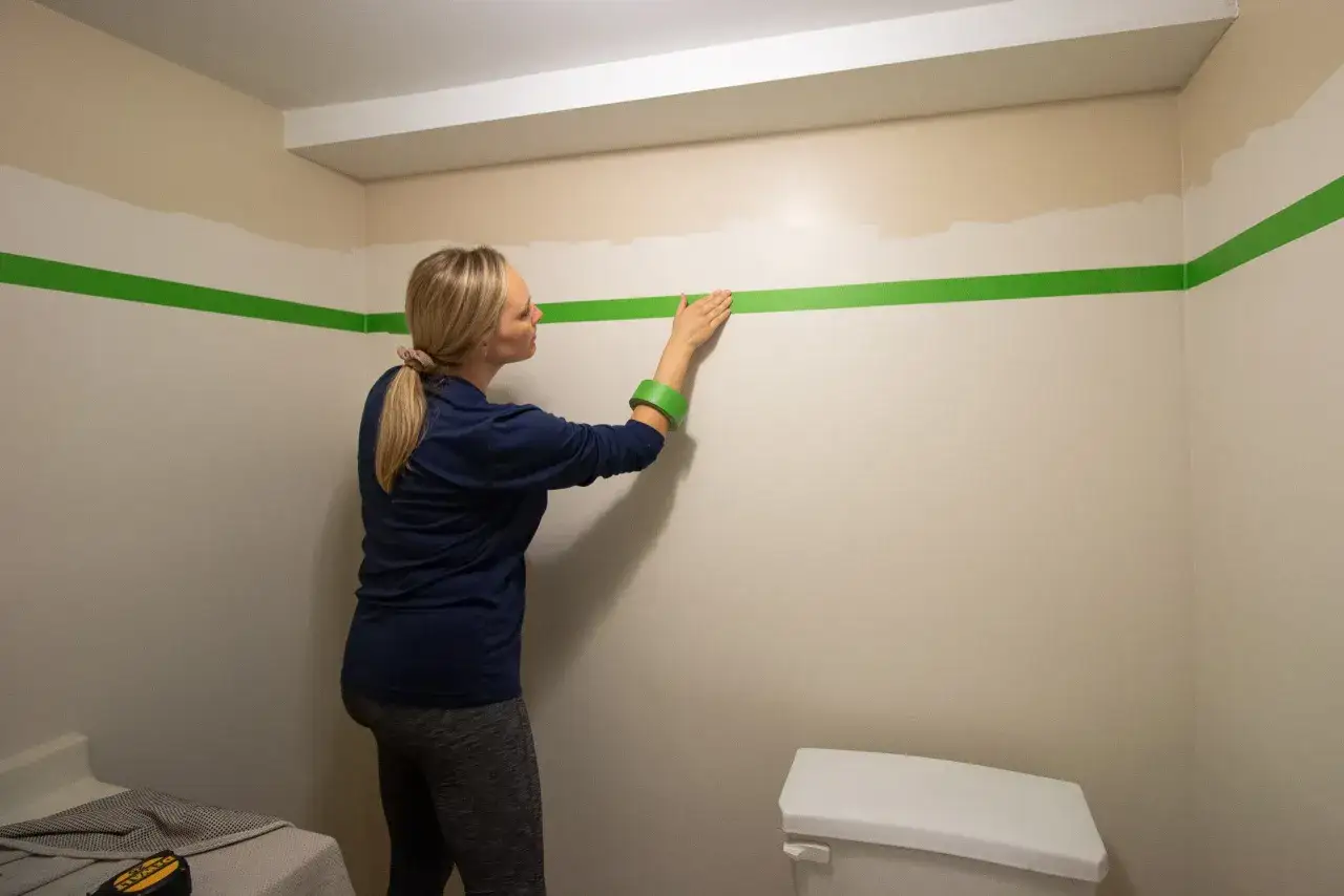 Une femme applique du ruban vert sur un mur pour créer une séparation entre deux couleurs.