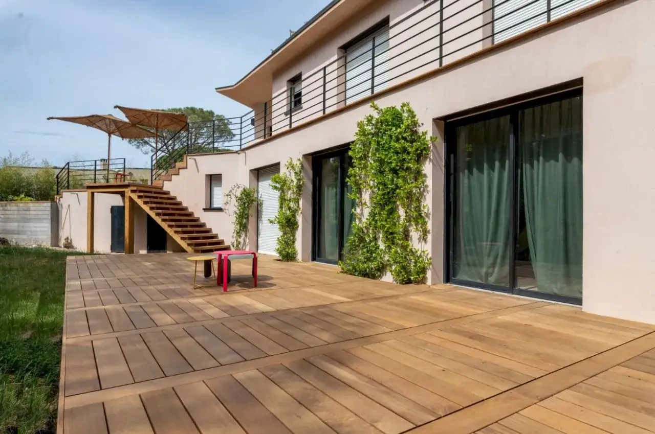 Une maison moderne avec les plus belles terrasses en bois s'ouvrant sur un jardin verdoyant. Des escaliers en bois m&egrave;nent &agrave; un niveau sup&eacute;rieur avec des parasols.