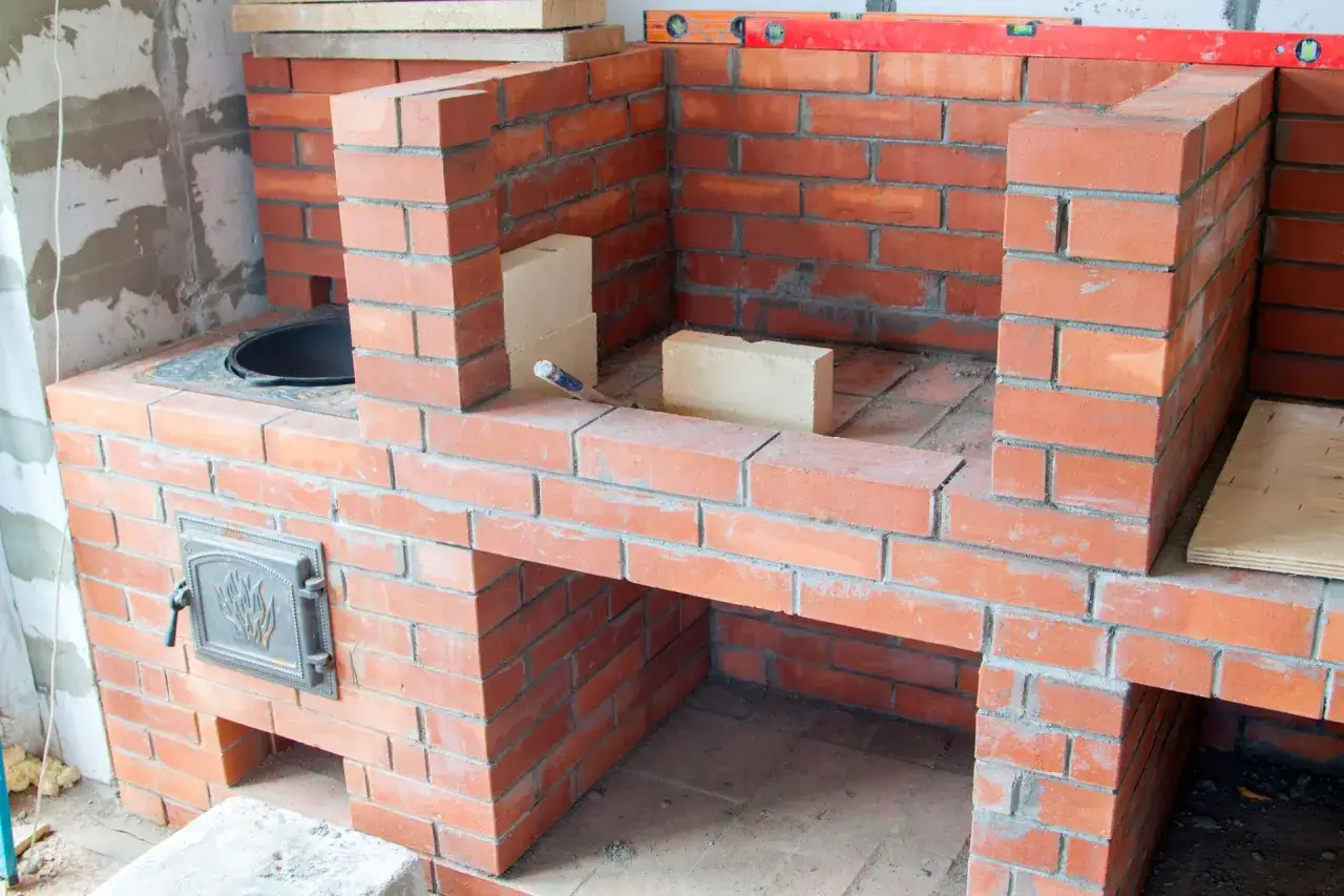 Construction d'un barbecue en brique, avec une porte en métal ornée et des briques claires pour la finition.