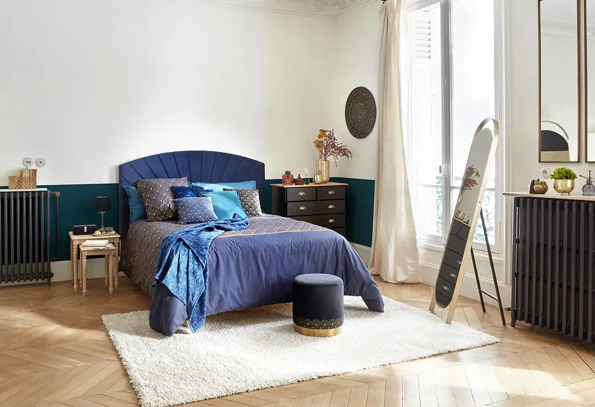 Chambre bleu nuit et doré, lit confortable avec coussins assortis, commode noire, miroir sur pied et tapis moelleux.
