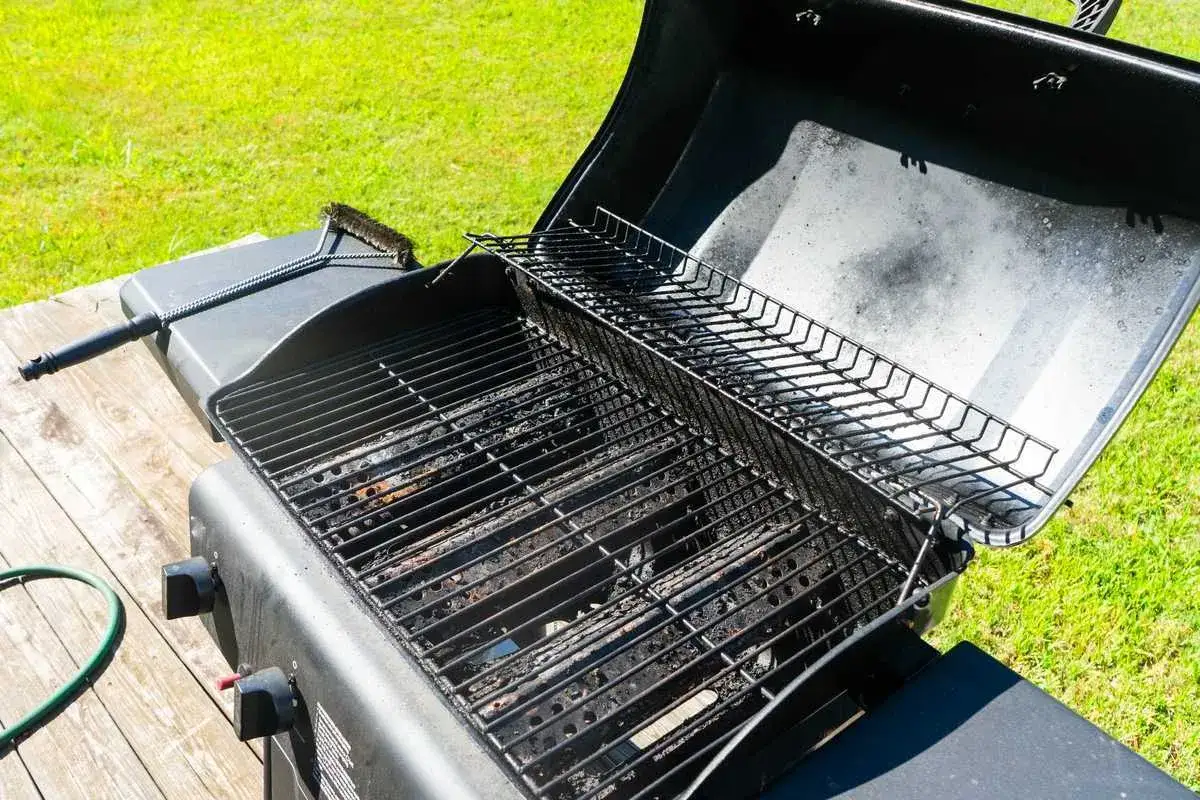 Un barbecue à gaz prêt à nettoyer, avec une brosse à portée de main. Les grilles sont sales, signe qu'il est temps de le faire briller pour la prochaine cuisson.