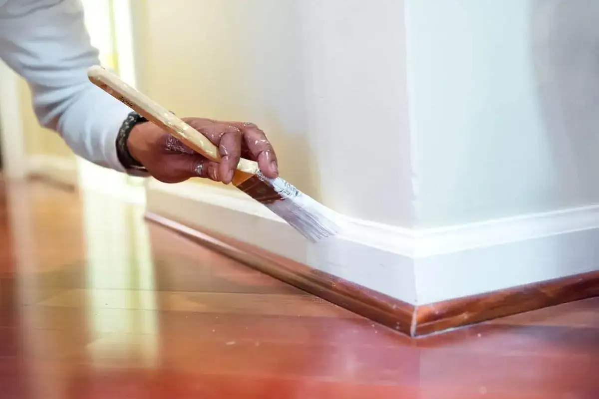 Main tenant peignant la plinthe blanche avec un pinceau, juste au-dessus du carrelage en bois.