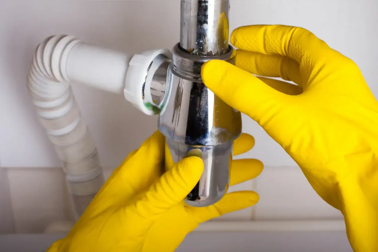 Mains jaunes pour changer le bonde du lavabo, manipulant le siphon chromé et le tuyau blanc.