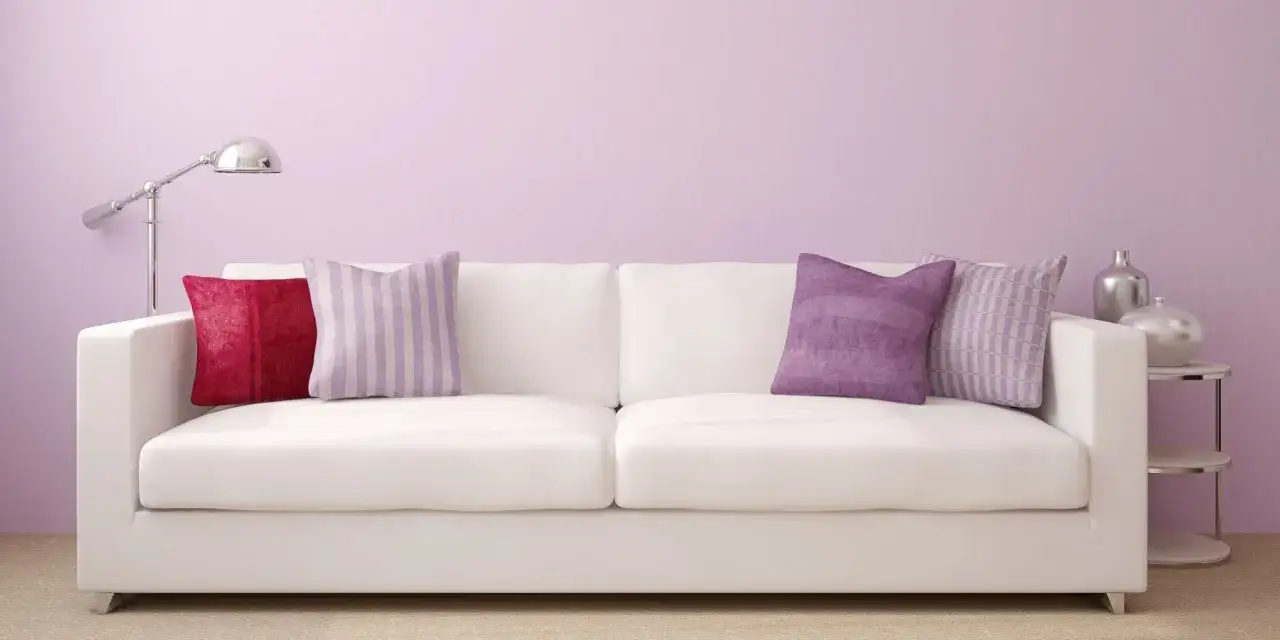 Canapé blanc avec coussins aux motifs variés, dont un à rayures dans un doux couleur parme clair, devant un mur pastel.