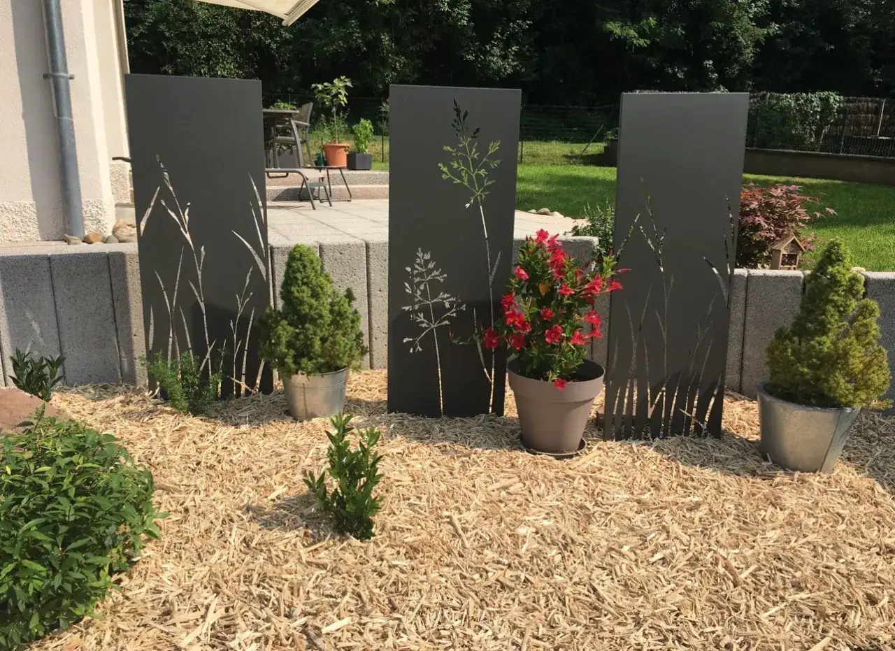 Panneaux décoratifs découpés avec des motifs de plantes, posés sur un lit de broyat de bois. Un pot de fleurs rouges et des conifères complètent la scène.