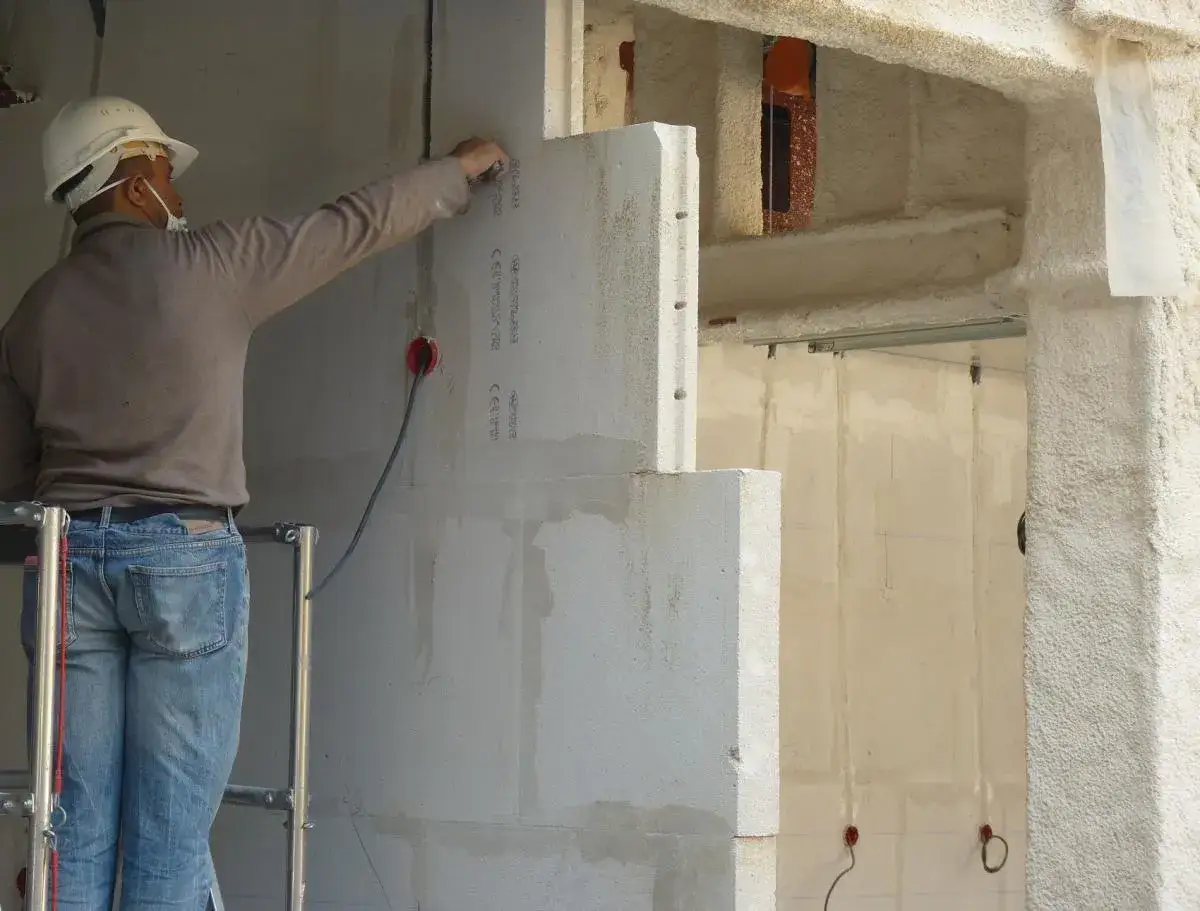Un ouvrier ajuste un bloc de béton cellulaire sur un mur en construction. Des fils électriques sont visibles.