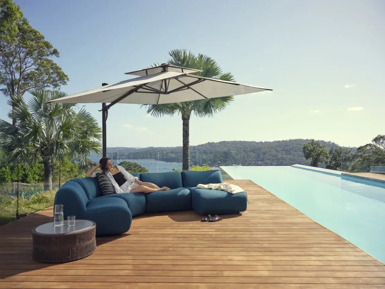 Femme d&eacute;tendue sur un canap&eacute; bleu, mobilier de jardin r&eacute;sistant aux intemp&eacute;ries, pr&egrave;s d'une piscine &agrave; d&eacute;bordement avec vue sur la mer.