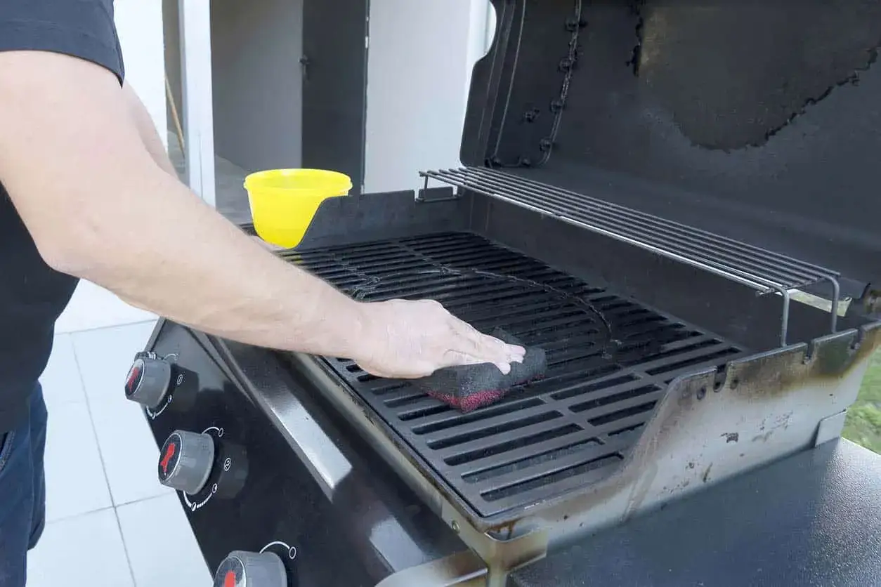 Un homme nettoie un barbecue très encrassé avec une éponge. Un seau jaune est à proximité.