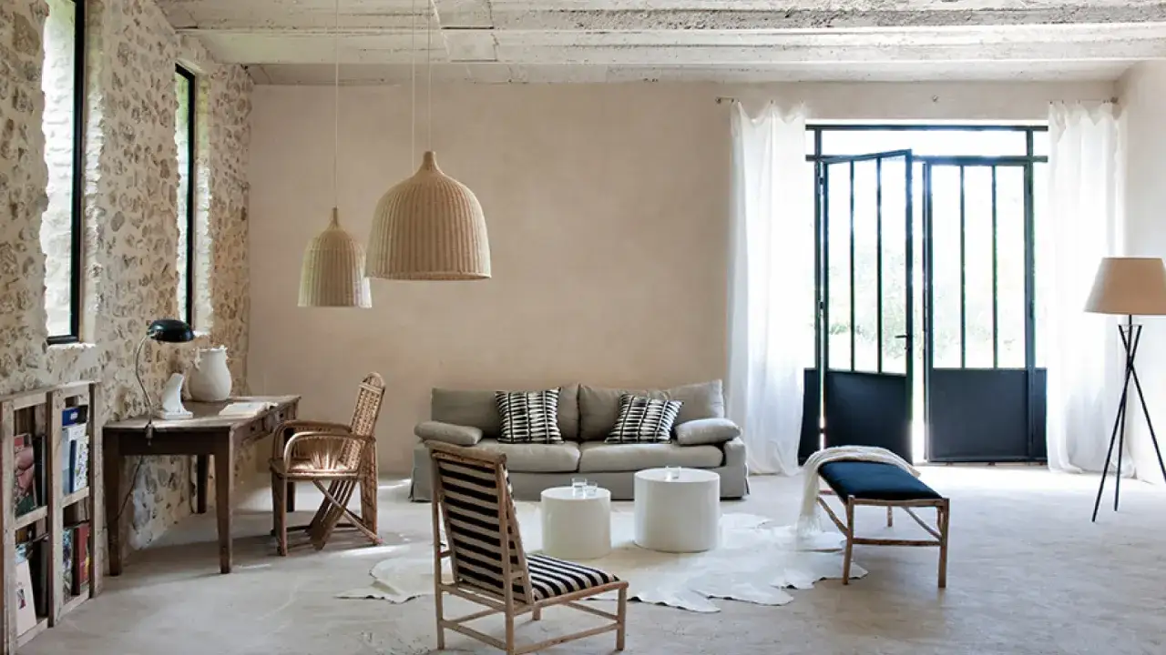 Salon lumineux avec murs en pierre et un bel effet enduit d&eacute;coratif. Meubles en rotin, canap&eacute; gris, et une grande porte vitr&eacute;e.