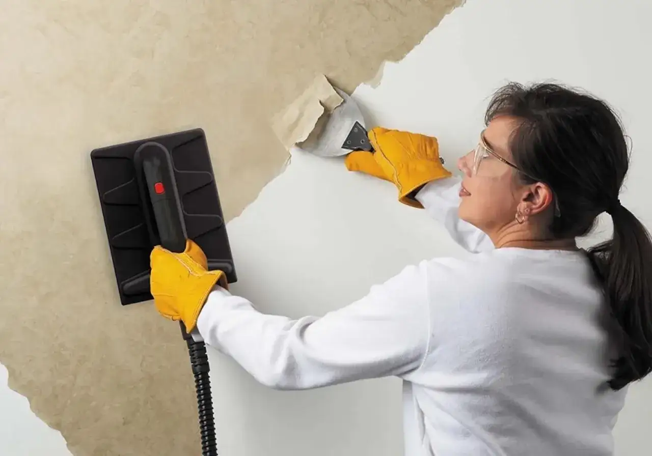 Une femme, portant des gants jaunes, utilise un appareil pour décoller du papier peint ancien d'un mur.