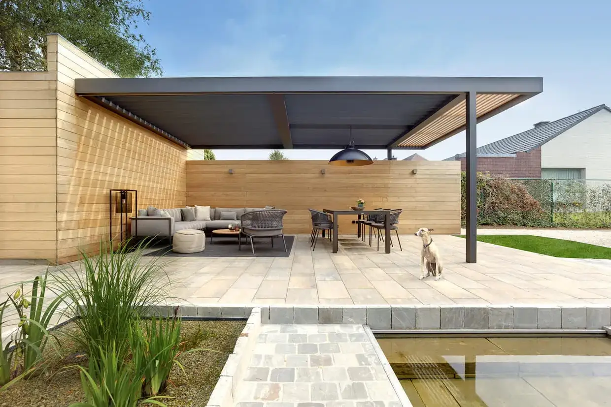 Une &eacute;l&eacute;gante terrasse en L avec salon et coin repas sous un auvent moderne. Un chien guette, profitant de la qui&eacute;tude.