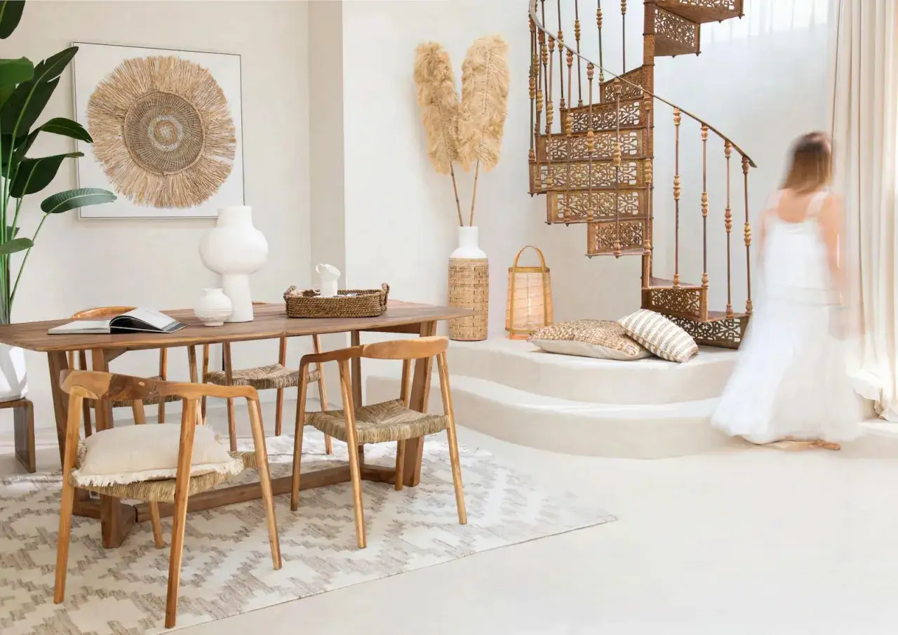 Un salon lumineux au style bohème chic maison, avec une table en bois, des chaises en rotin, un tapis graphique et un escalier en colimaçon.
