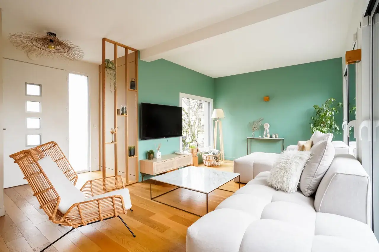 Salon lumineux aux murs d'un nuancier de vert tendance, avec canap&eacute; blanc, fauteuils en rotin et mobilier en bois.
