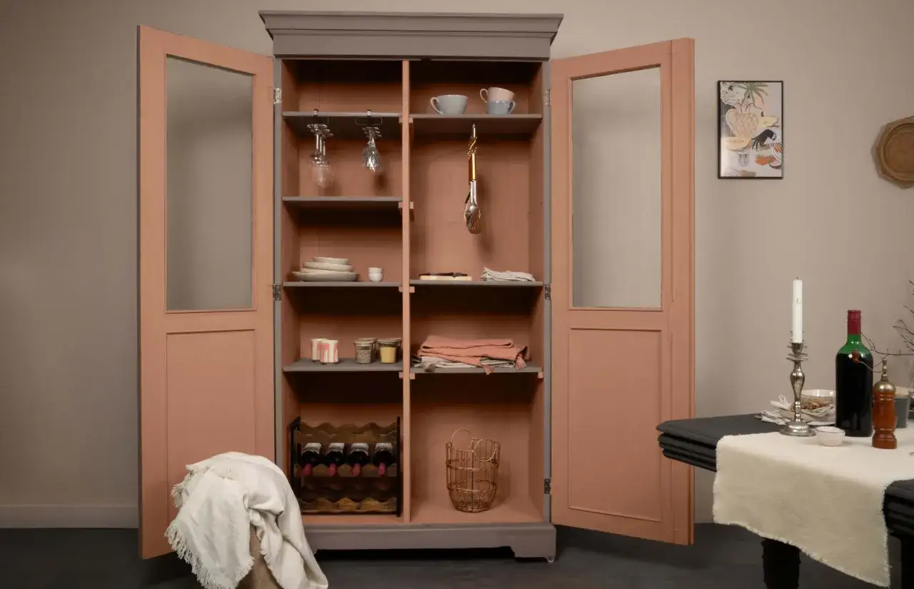 Armoire vitrée avec un relooking meuble 2 couleurs : gris anthracite et rose poudré. Elle contient vaisselle, verres, bouteilles de vin et objets déco.