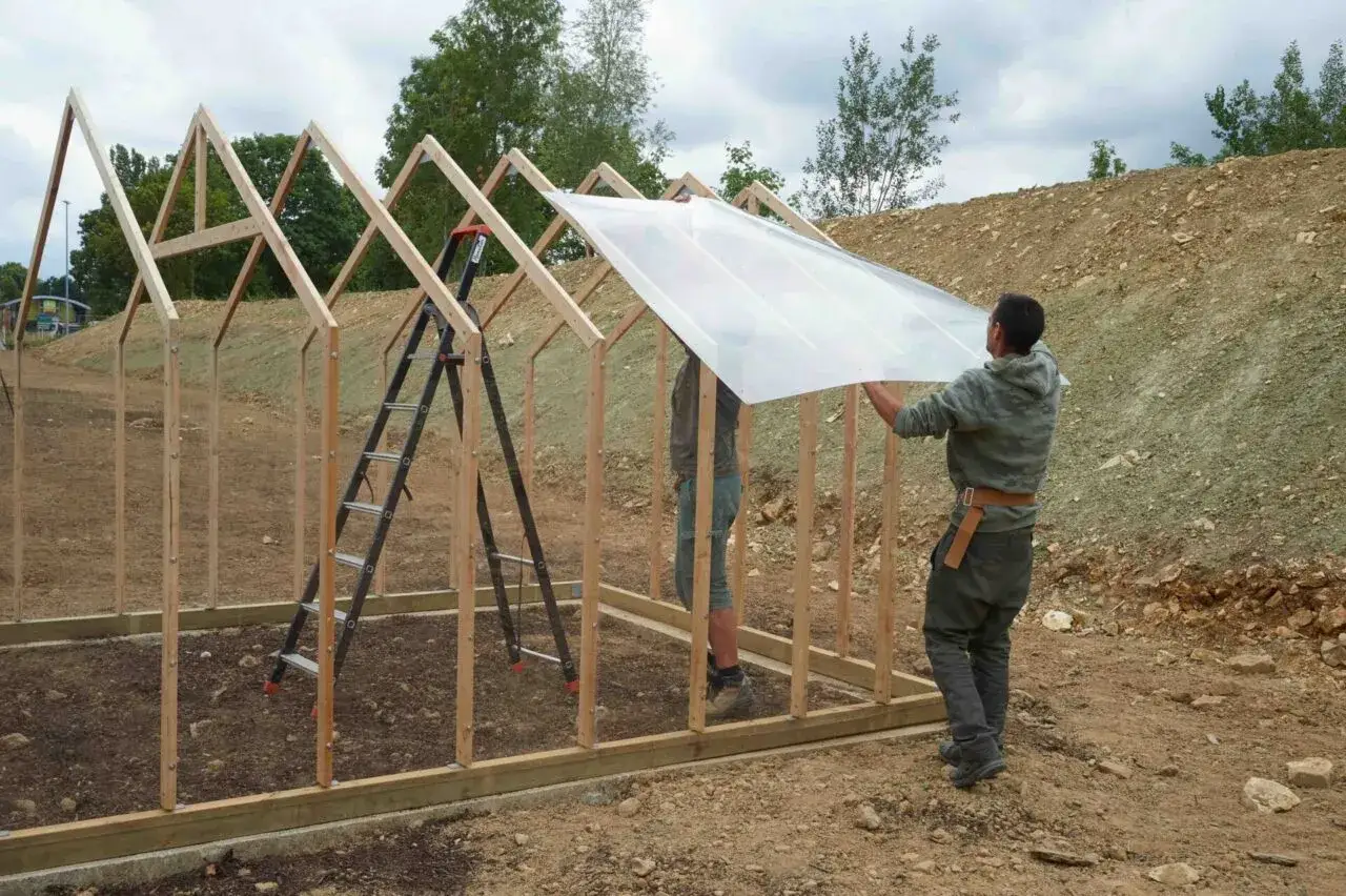Deux hommes travaillent à fabriquer une serre en bois, posant une grande plaque de plastique transparent sur la structure.