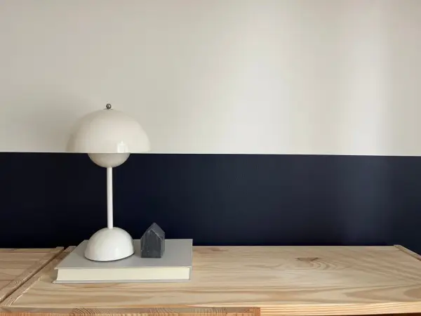 Lampe blanche sur un livre, devant un mur deux couleurs différentes, blanc et bleu nuit. Un petit objet gris en forme de maison est posé sur le livre.
