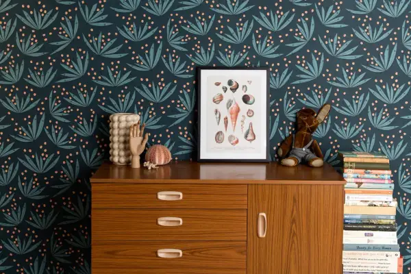 Idée motif peinture mur : papier peint botanique bleu nuit avec des feuilles stylisées et des baies roses. Un cadre de coquillages et une commode vintage complètent la scène.