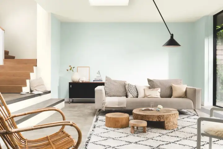 Salon lumineux aux murs couleur vert d'eau, avec un canapé beige, des tables basses en bois brut et un fauteuil en rotin.