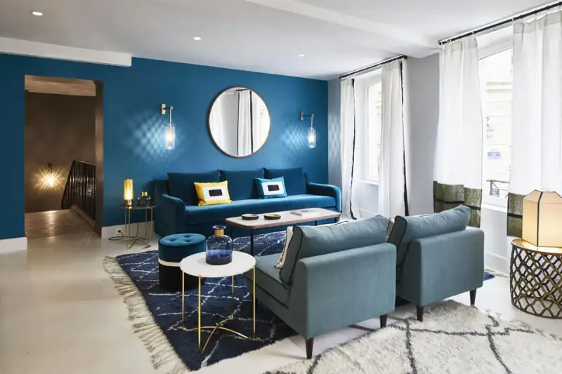 Salon moderne aux murs bleu canard, avec canapé velours, fauteuils assortis, tapis graphique et éclairage d'ambiance.